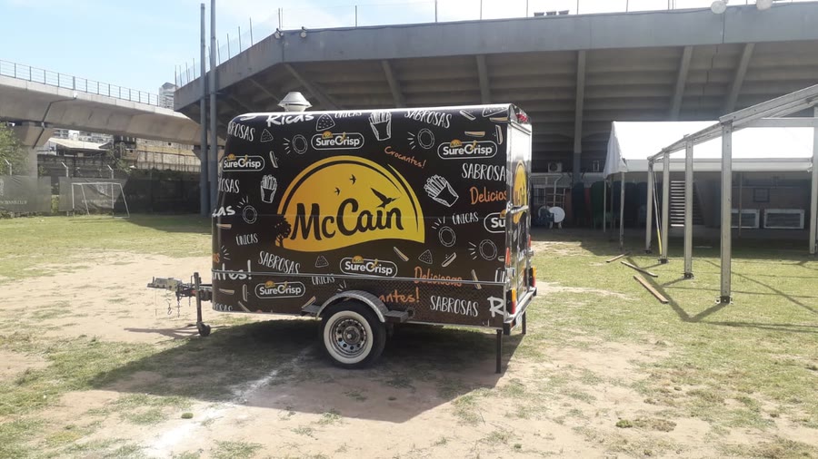 Trailer de OCHO ploteado con la gráfica de McCain, sobre el césped de un predio con tribuna